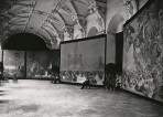 4 canvases are exhibited in a large hall with an ornate cieling and a wooden floor; a man sits on a stool in front of one of the canvases