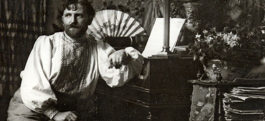 Mucha in his studio dressed in an embroidered smock, seated and leaning on a piece of furniture