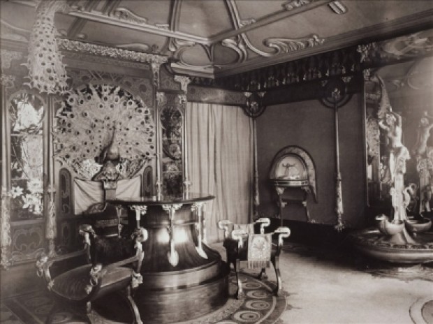 A black and white general view of the boutique interior with a peacock decoration on the far wall, a wooden circular counter with 2 elaborate stools and a presentation case and water feature on the right wall