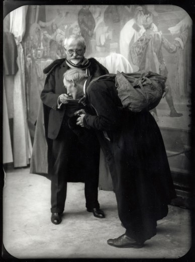 Dressed in a suit and a cape, Mucha holds out a dish for an elderly man with a long coat and a rucksack who bends over and touches the dish with his face