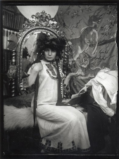 Model in a white embroidered tunic and a black frilly headpiece sits on a wite rug on a couch in front of an ornate gilt mirror