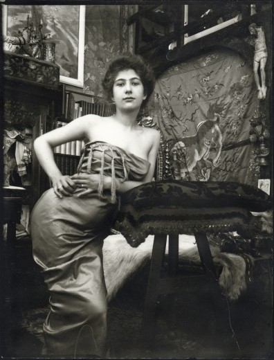 Young bare-shouldered model wrapped in a large piece of fabric leans on a cushion on a pedestal with her left elbow and holds her right hand on her right hip