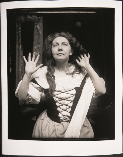 A Woman in a white dress with a laced up black bodice holds her hands out beside her face and looks upwards