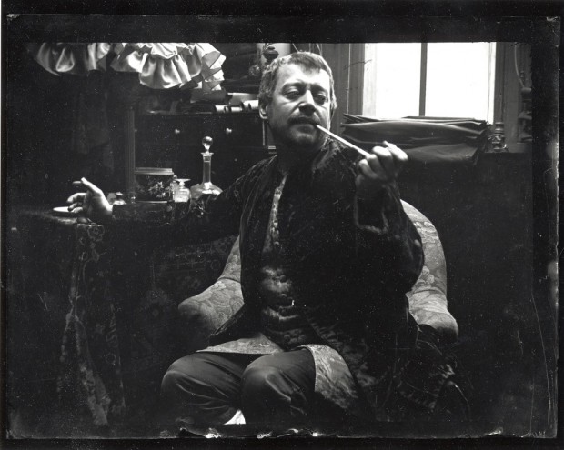 Seated model in a velvet smoking jacket holding a pipe to his mouth and reaching out with his right arm to a table behind him