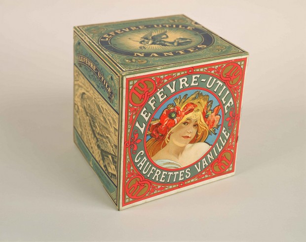 A square box seen from the side with a blond woman with poppies in her hair in a red circular form with the words 'Lefèvre-Utile Gaufrettes Vanille' on one side, and a picture of the LU factory in black and white on one of the other sides.