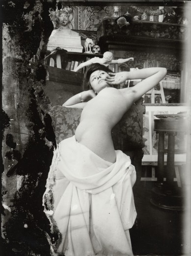 Nude female model with a white sheet draped around her lower body leaning back over a chair with her arms raised to shoulder level and her hands to her face
