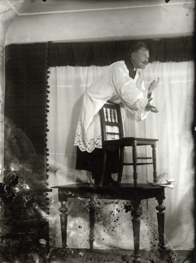 Dressed as a priest, Mucha stands on a table and leans forward over the back of a chair placed on the table