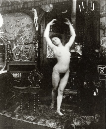 Nude in fifth position balancing on her left leg holding her right foot just behind her with both arms arched above her head