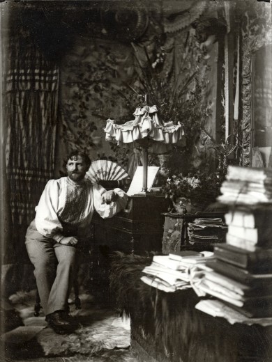Mucha seated in his studio with ornate fabric in the background, art objects to his right, and piles of books in the foreground