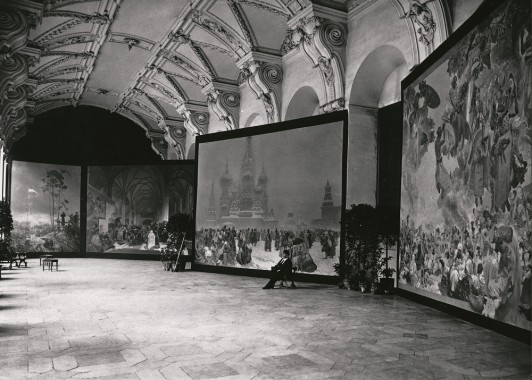 Klementinum exhibition, Prague 4 canvases are exhibited in a large hall with an ornate cieling and a wooden floor; a man sits on a stool in front of one of the canvases