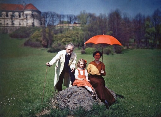 Mucha, Maruška and Jaroslava in the grounds of Zbiroh Castle Maruška and Jaroslava sit on a rock with Mucha standing to their left in a green field with a wing of the castle visible in the top left of the photo; Maruška holds a red parasol and Mucha holds a walking stick