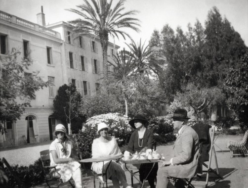 Four figures sat around a table in a garden with palm trees in front of a large building