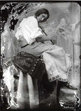 Girl in a voluminous white shirt and a traditional skirt holding a broom handle sitting on an ornate rug in front of one Mucha's canvases