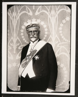 Self-portrait in formal masonic regalia Mucha dressed in formal wear with a cap, a sash, a medallion and a medal, stood in front of a panel decorated with stars and arabesques