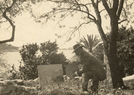 Mucha painting in Croatia Mucha sits next to an easel amid trees on a coastline with a view of a bay