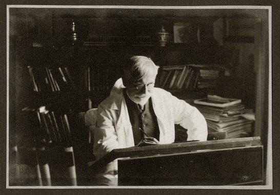 Mucha working Mucha sitting at a drawing table with a bookshelf behind
