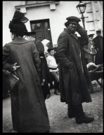 Woman seen from behind wearing a hat, a long coat and a handbag stood next to a beggar with his left hand to his cap and a frown on his face