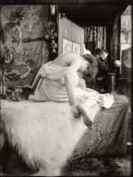 Bare-shouldered model sits on a white rug on a couch and leans forward away from the camera