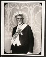 Mucha dressed in formal wear with a cap, a sash, a medallion and a medal, stood in front of a panel decorated with stars and arabesques