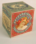 A square box seen from the side with a blond woman with poppies in her hair in a red circular form with the words 'Lefèvre-Utile Gaufrettes Vanille' on one side, and a picture of the LU factory in black and white on one of the other sides.