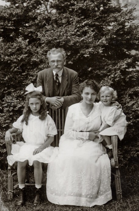 Maruška, Jaroslava and Jiří sit on a bench in front of a bush while Mucha leans on the bench behind them