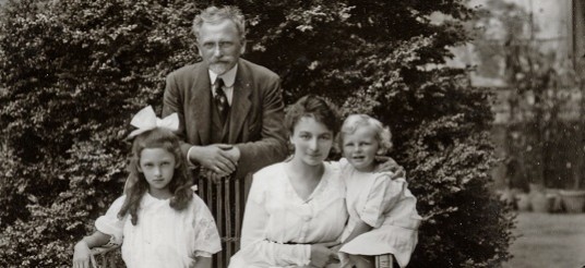 Maruška, Jaroslava and Jiří sit on a bench in front of a bush while Mucha leans on the bench behind them