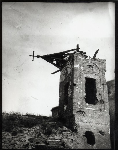 A ruined church tower stands against a grey sky with its wooden steeple toppling over to the left