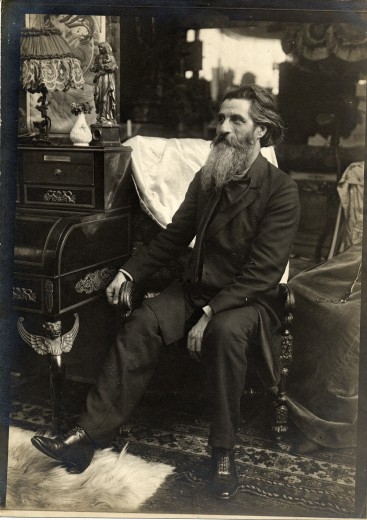 A man with dark hair, a long grey beard and a dark suit sits next to a desk looking out to the right of the camera