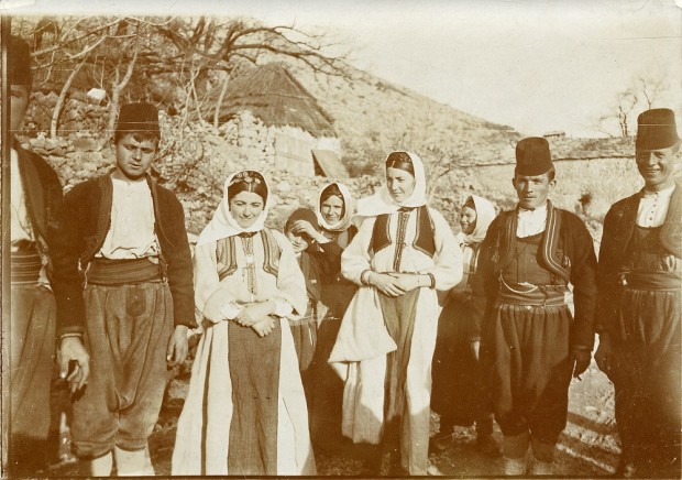 A group of four men, four women, and a small boy stand in a line in front of a stone settlement