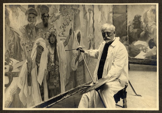 Mucha sits in overalls on a stool in front of his canvas holding a paintbrush in his right hand and a palette in his left hand; he looks out at the camera