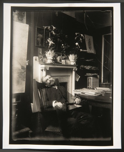 Mucha sitting back on a chair next to a table and in front of a well-adorned fireplace