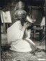 A female model dressed in a white tunic with fabric in her long, dark hair kneels on the carpeted floor with her face in her hands