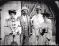 Four figures dressed in formal dress and hats standing in front of a wall and a wooden gate