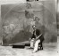 Mucha seated on a stool in front of his mural with a paint palette and paint brush in his hand